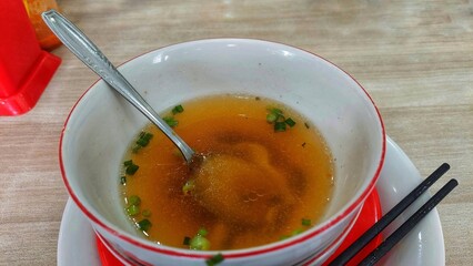 A close-up of a bowl of clear broth with chopped green onions, a spoon, and chopsticks.
