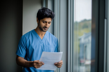 Male doctor reading a report while standing by the window indoors  