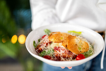 Fresh Caesar salad with croutons, cherry tomatoes, grated cheese, and bacon in a white bowl, held outdoors by a person in white shirt, blurred background.
