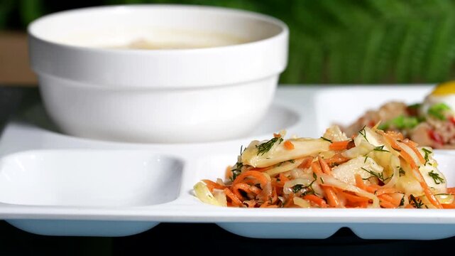 Close-up of fresh vegetable salad with carrots, cabbage, dill, next to a white bowl of creamy soup on a white tray.