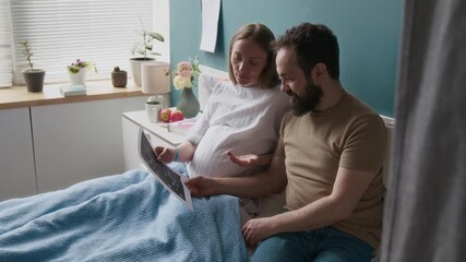 Medium full shot of cheerful pregnant young Caucasian woman demonstrating baby ultrasound photo to happy husband, chatting, joking and laughing. Supportive man visiting wife in maternity clinic - Powered by Adobe