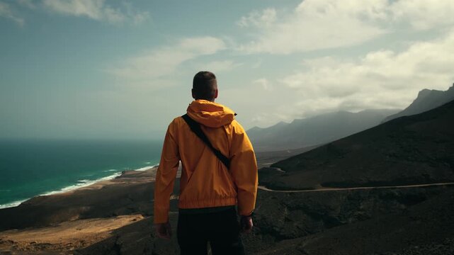 Man in orange jacket facing scenic view of Cofete beach from mountain viewpoint, strong wind blowing, Fuerteventura, Canary Islands, travel concept slow zoom out