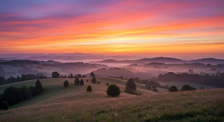Naklejka premium A vibrant sunrise paints the sky with orange and pink hues over a misty, rolling landscape of hills and scattered trees.