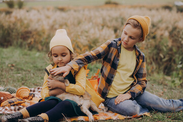 Little cute smiling girl and boy sitting on plaid with cat. Kid resting with her family in the nature. Picnic. Autumn season.