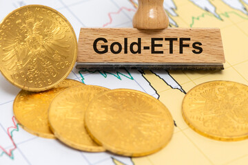 Ein Holzstempel ist beschriftet mit Gold-ETFs über echten Goldmünzen und Börsenkurs