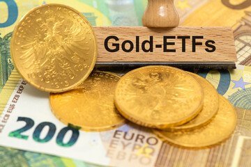 Ein Holzstempel ist beschriftet mit Gold-ETFs über echten Goldmünzen und Euro Banknoten