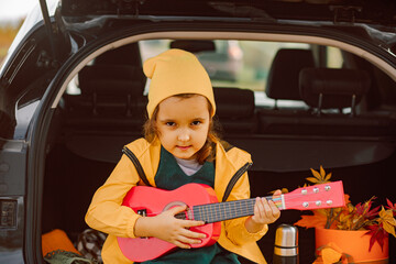 Little cute smiling girl sitting near by on open car trunk. Girl playing small guitar. Kid resting with her family in the nature. Autumn season.