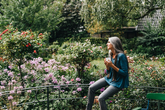Woman with coffee mug sitting on railing of balcony looking at distance
