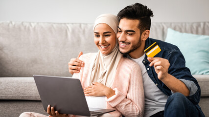 Happy expectant muslim couple shopping online with laptop and credit card at home while relaxing...