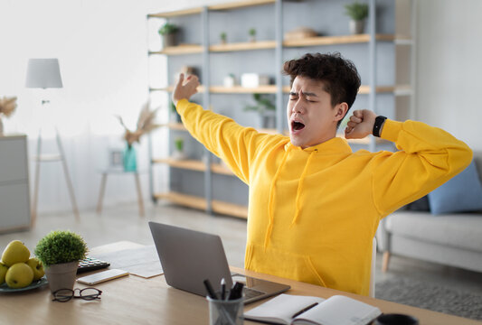 Tired asian male student yawning sitting at desk using laptop. Bored guy is exhausted from getting ready for test or writing coursework, feeling sleepy, stretching arms. Lack of sleep and tiredness