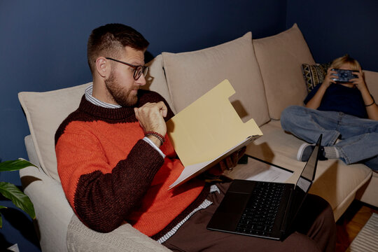 Father reading a document on a sofa while son uses a smartphone at home