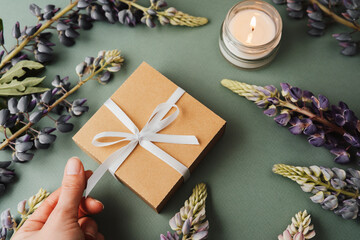 Hand holding gift box with candle and lupine flowers on green background