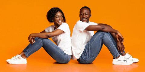 Carefree african american lovers sitting back to back on floor, looking at each other and smiling on orange background, studio shot, copy space. Cheerful millennial black man and woman posing together