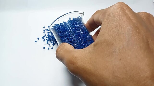 Pouring blue masterbatch granules with glass container, polymer carrying blue pigment is poured in white background, suitable for industrial plastic company profile videography catalog design.