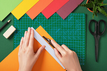 Hands cutting colored paper with utility knife on green cutting mat in top view. Creative craft process, handmade project preparation.