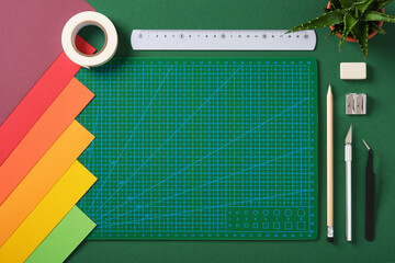 Crafting tools and colored paper on green background with empty cutting mat. DIY, hobby, scrapbooking and creative project preparation.