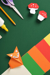 Origami fox and mushrooms with craft supplies on green paper background. Concept of creative kids crafts and handmade paper projects.