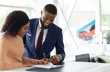 Happy Black Woman Buying New Car, Signing Papers With Male Salesman In Dealership Office, Cheerful African American Female Driver Purchasing New Automobile In Modern Showroom, Copy Space