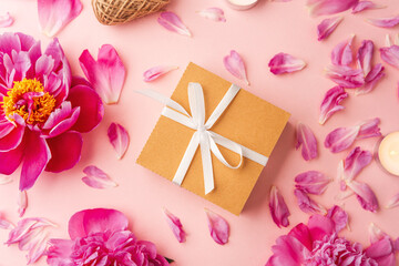 Gift box with white ribbon surrounded by peonies, petals, candles and twine on pink background