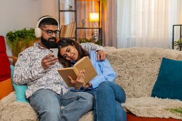 Indian couple man and woman sitting on home sofa, spending calm day together as girl reads book and...