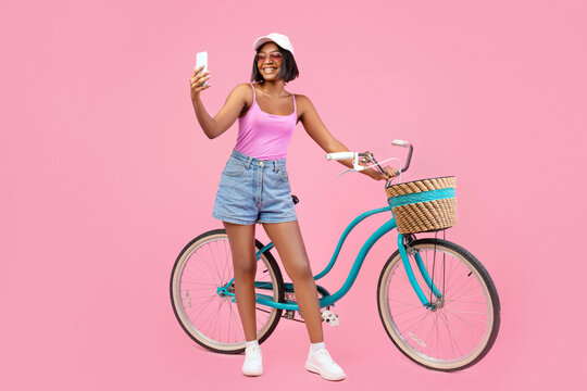 Full length of cheerful black lady in summer clothes taking selfie on smartphone next to classic bicycle on pink background. Lovely young woman making photo of herself near bike. Summertime sports