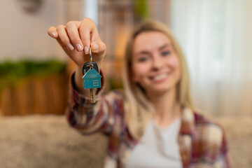 Young smiling adult woman holds new keys while sitting on home sofa, proudly celebrating apartment...