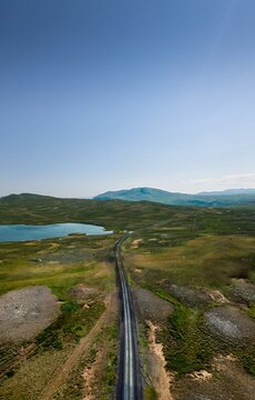 Aerial view of a dark road cutting through a vibrant green and brown landscape, leading towards distant mountains and a serene blue lake, Raufarh&Atilde;&para;fn, Nor&Atilde;&deg;ur&Atilde;&frac34;ing, Iceland.