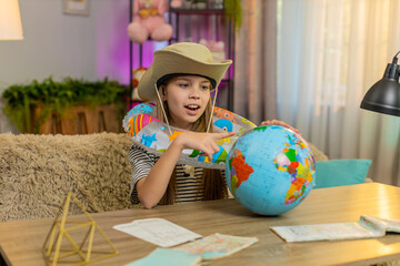 Happy child girl sitting at home table wears a travel hat and inflatable ring while looking at a...