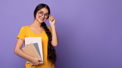 Education Concept. Portrait of happy indian female student wearing and touching eyeglasses, holding notebooks looking at camera standing isolated over purple studio background wall with free space