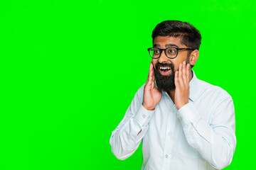 Indian man lifts eyebrows and opens eyes wide in amazement, placing hands near face, reacting to...
