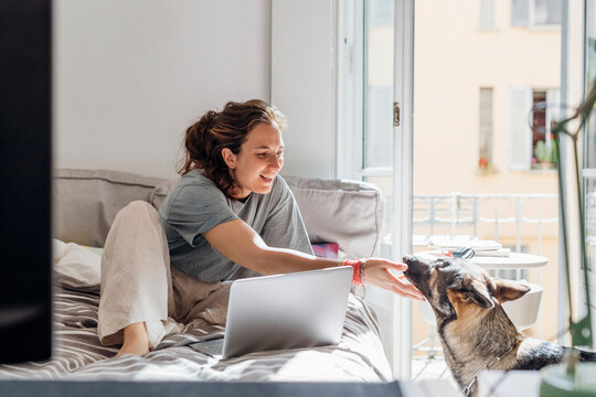 Happy female freelancer touching dog while sitting on bed