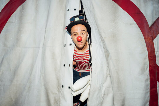 Male clown with hat and juggling pin peeking through circus tent entrance
