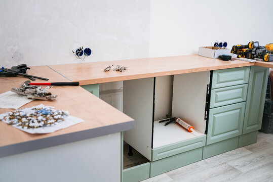 Carpenters are assembling a new kitchen in an apartment, installing cabinets, countertops, and various tools and materials are scattered around the workspace