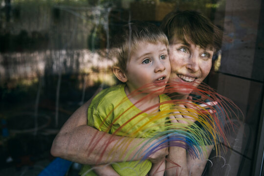 Portrait of happy mother and her little son looking out of window with drawn rainbow