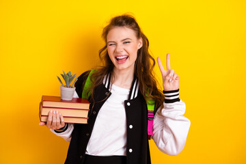 Cheerful student girl showing peace sign with book against bright yellow background reflecting...