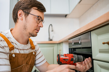 Skilled handyman wearing overalls and safety glasses carefully installs a new oven in a modern kitchen using a cordless drill, ensuring precise and secure placement
