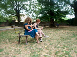 Two young women sitting on a park bench together engrossed in their smartphones in a green outdoor...