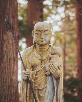 View of a serene stone Jizo statue holding a baby, bathed in soft light amidst the blurred trees, a peaceful scene, Gyokusenji Temple, Tsuruoka, Yamagata, Japan.