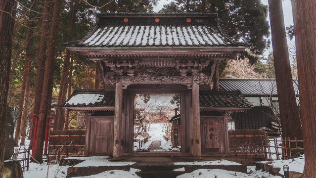 View of a weathered, ornate wooden gate stands amidst snow-dusted trees, leading to a temple bathed in soft light, Gyokusenji Temple, Tsuruoka, Yamagata, Japan.