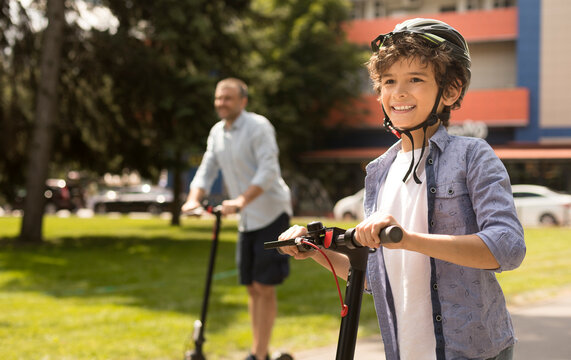 Portrait of happy curly boy in protective helmet having ride on motorized scooter with father in blurred background - Powered by Adobe