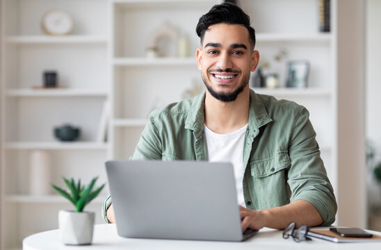 Glad young handsome muslim male with beard looking at camera, working on laptop in home office interior. Freelance, business remotely to workplace with device during covid-19 quarantine, free space - Powered by Adobe