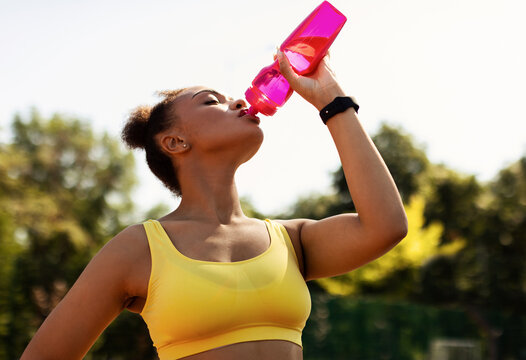 Sports And Hydration Concept. Portrait of fit black woman drinking fresh water from bottle, wearing yellow sportswear top bra, exercising oudoors taking break. Street Workout, Outside Training - Powered by Adobe