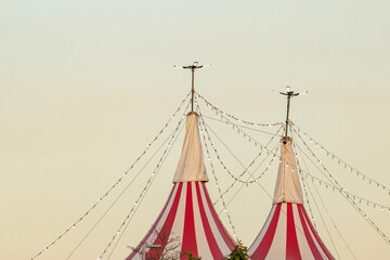 Topo da estrutura de um circo, com lonas listradas de vermelho e branco e varais de luzes acesas,...