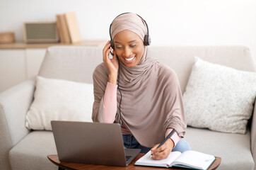 Joyful African American woman in hijab and headphones taking notes during online business meeting...