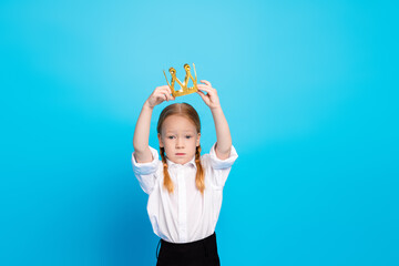 Young girl placing a golden crown on her head against a bright blue background, expressing...
