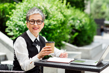 Mature professional woman with short hair working outdoors with a notebook and laptop while...