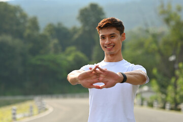 A smiling man preparing for fitness activity by stretching his hands on a quiet road with trees in...