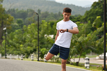A Fit man stretching his leg and checking his smartwatch before a run on a scenic outdoor road...