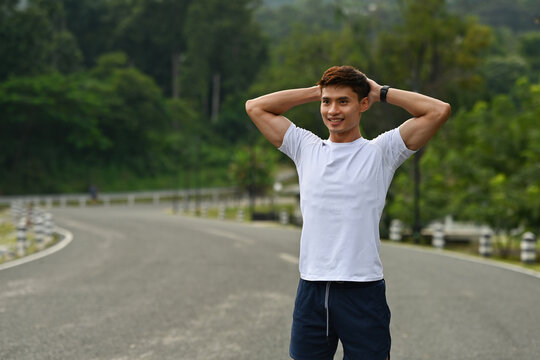 A Sporty man in white shirt doing pre-workout stretching outdoors in a green, natural environment