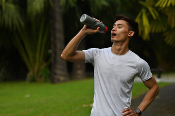 A young Asian man in sportswear drinking water from a bottle after a workout session outdoors, showing hydration and recovery